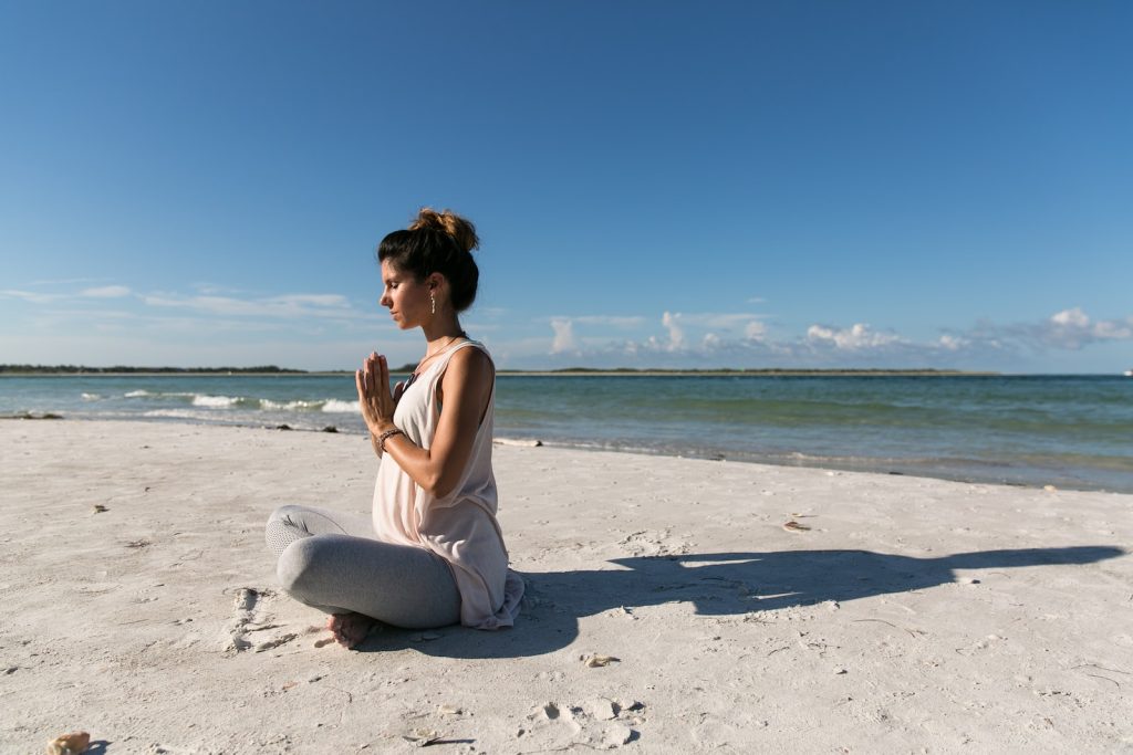 woman praying and sitting on sand seashore during day