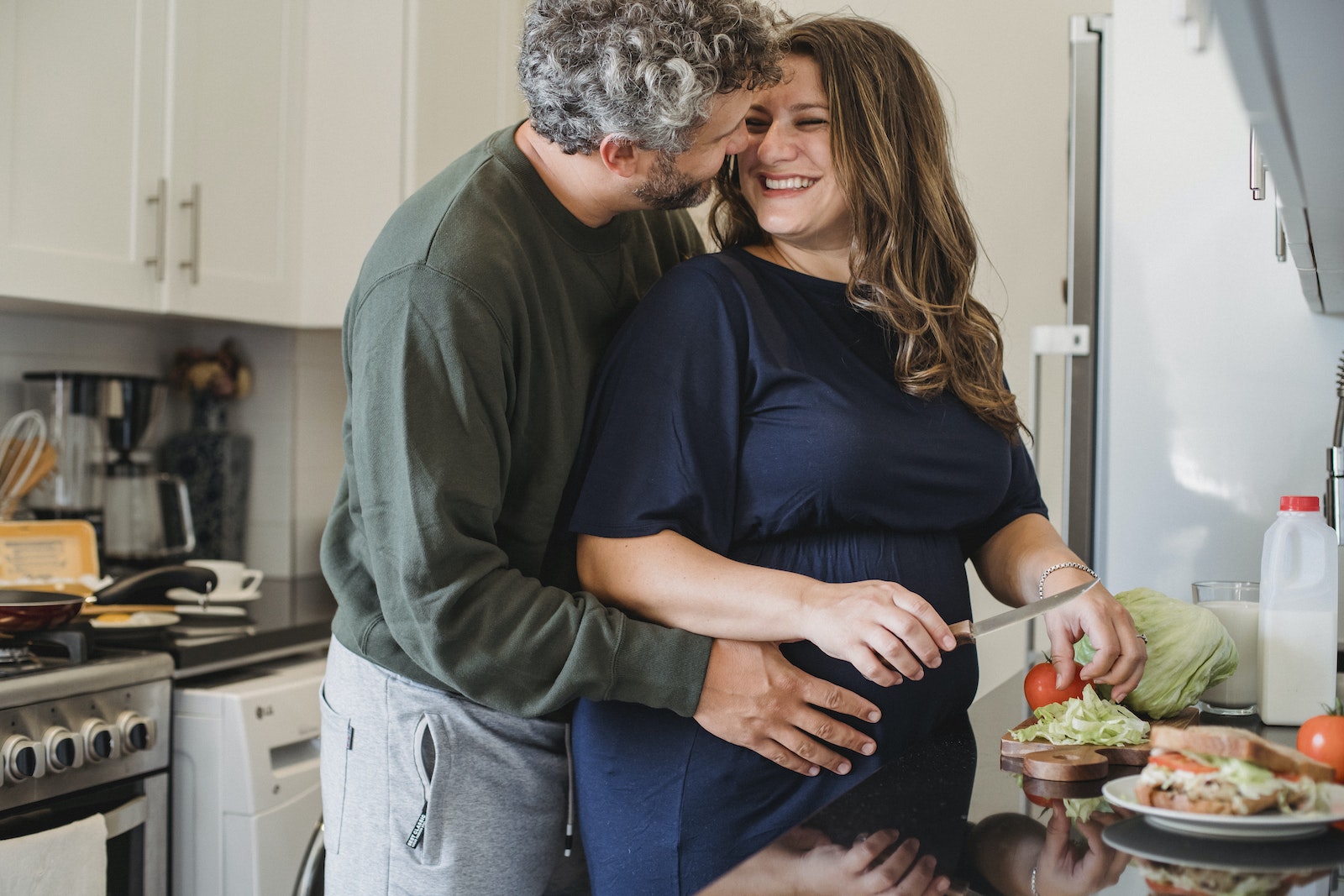 Cheerful pregnant wife in blue dress cooking sandwiches for breakfast while smiling and looking at husband embracing belly in modern kitchen